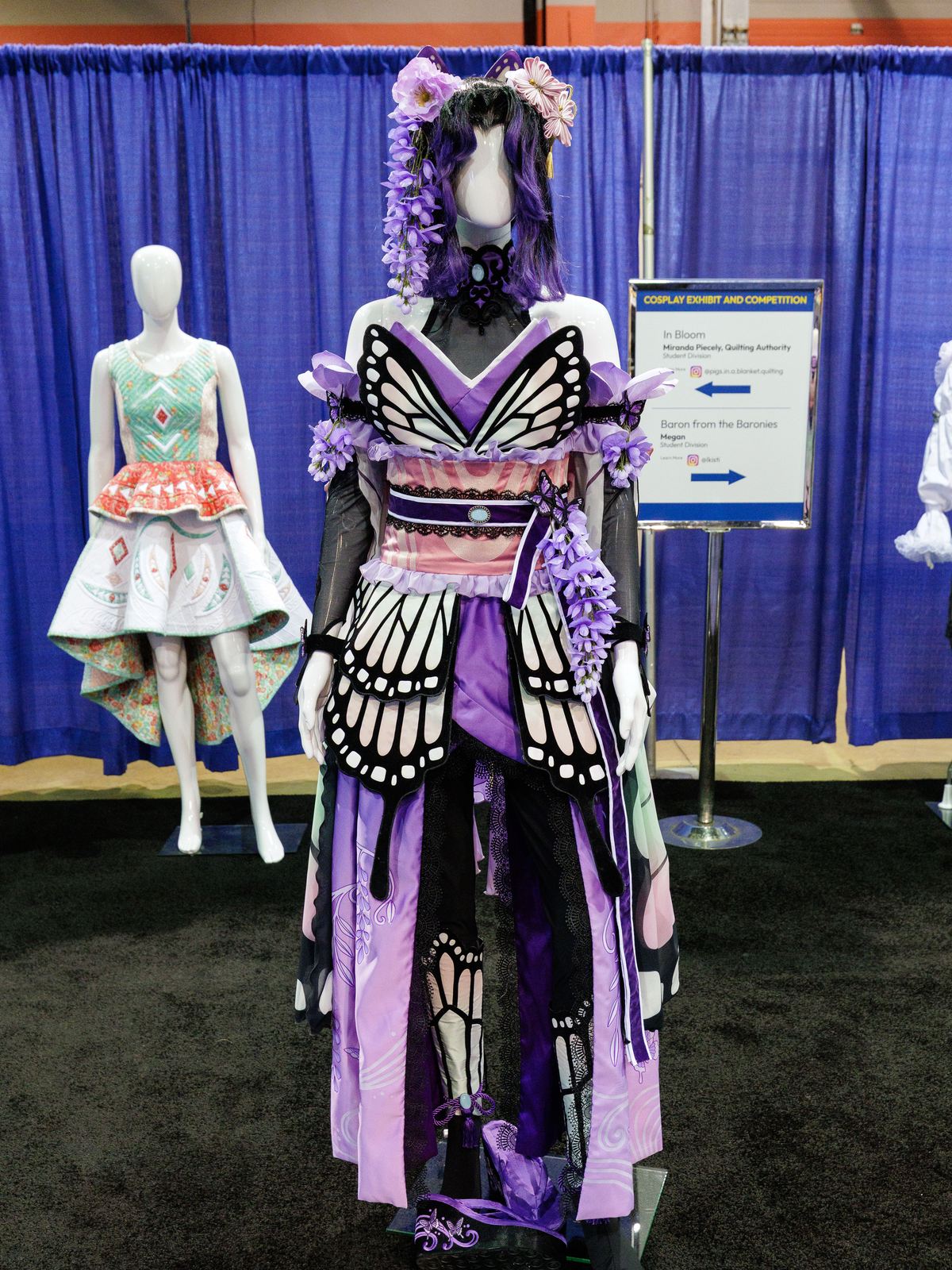 A cosplay costume on a mannequin: a purple kimono-style gown with black and white butterfly-wing panels and wisteria in the hair, shown at the h+h americas cosplay exhibit