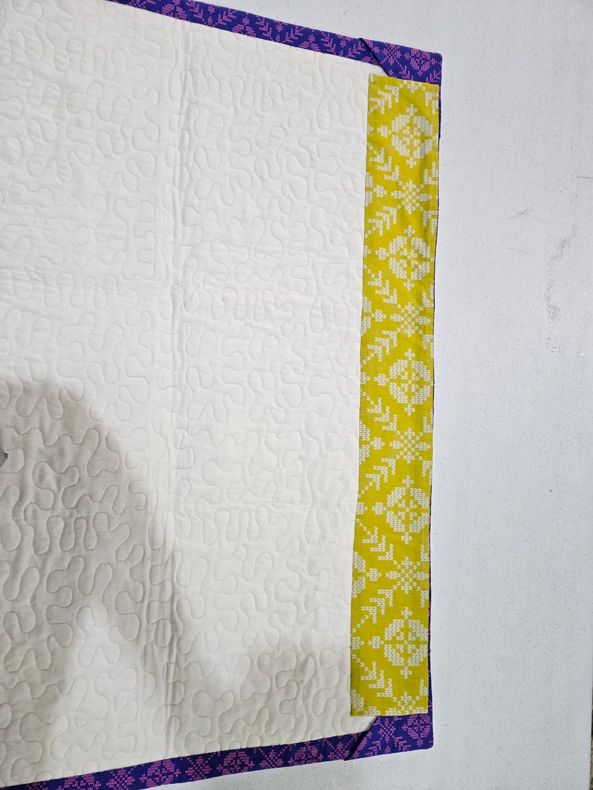 Close view of the quilting on a Loter&iacute;a piece: dense stipple stitching across the cream backing, bordered by the yellow strip and purple binding