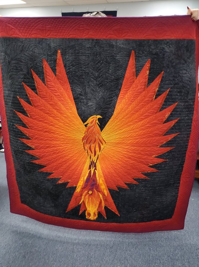 A quilt of a phoenix rising with wings spread, in oranges and reds against a grey ground with a deep red border